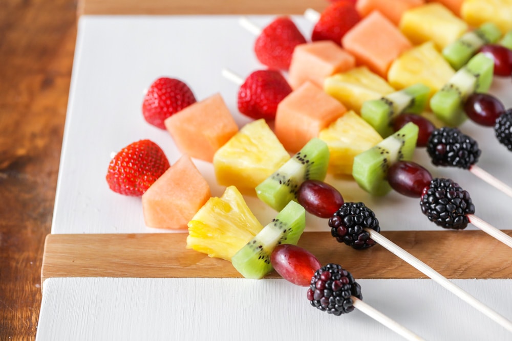 简单的开胃菜——草莓、哈密瓜、菠萝、猕猴桃、葡萄和黑莓串在木制串上，放在白色砧板上。