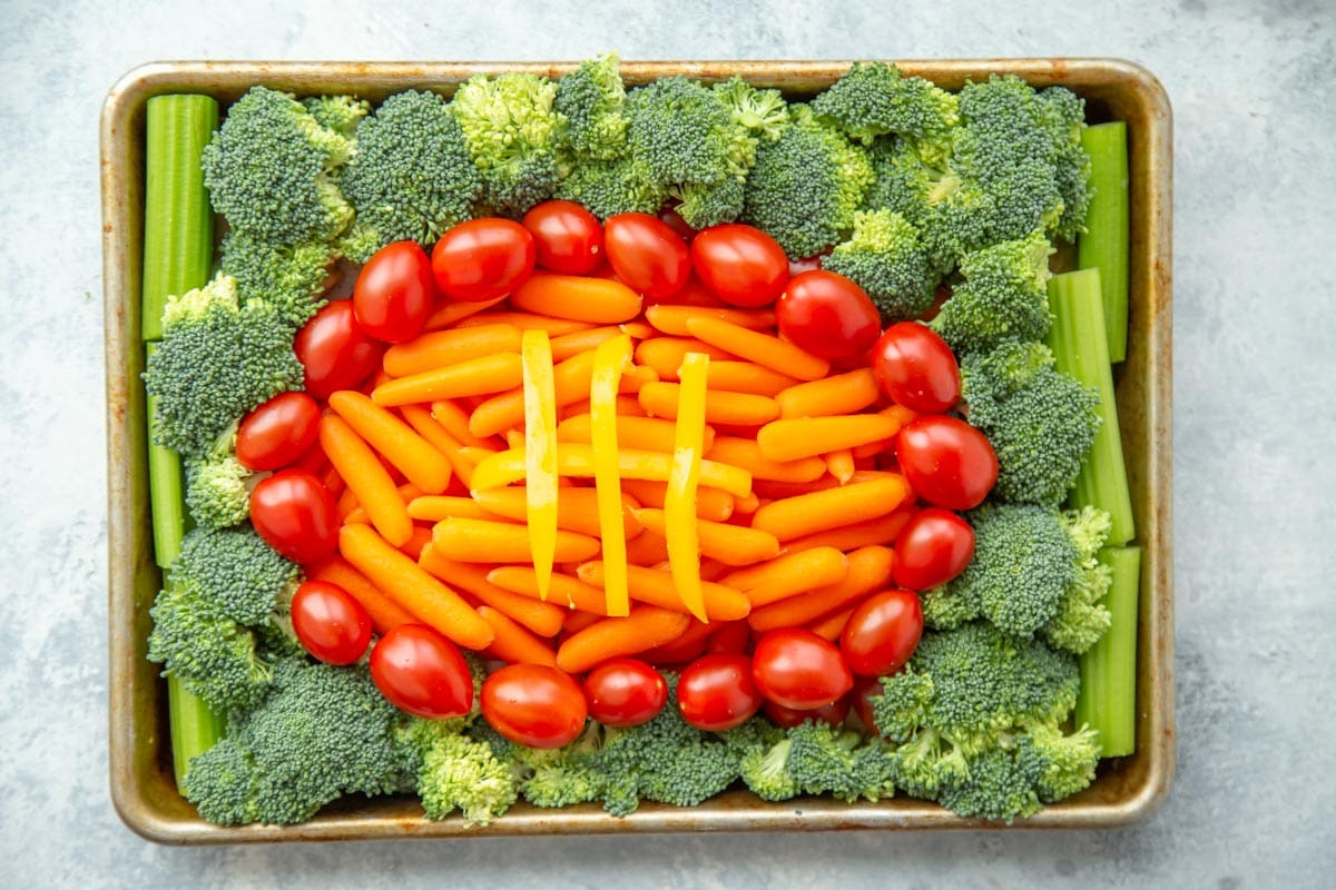健康的开胃菜——一个蔬菜托盘，上面放着小胡萝卜和葡萄番茄，看起来像一个足球，上面放着黄辣椒片，周围放着西兰花，放在烤盘上。