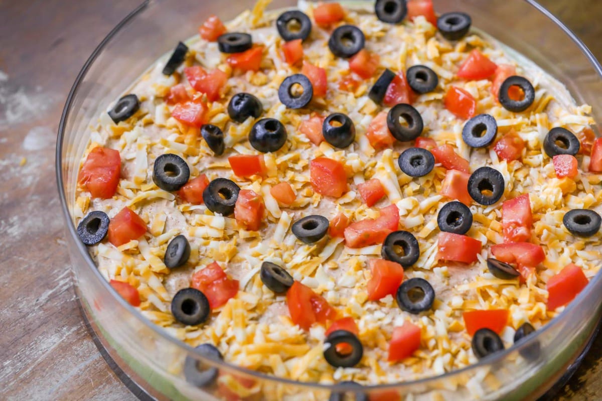 超级碗开胃菜- 7层豆浸在一个透明的玻璃碗上，上面有切片黑橄榄和西红柿丁。